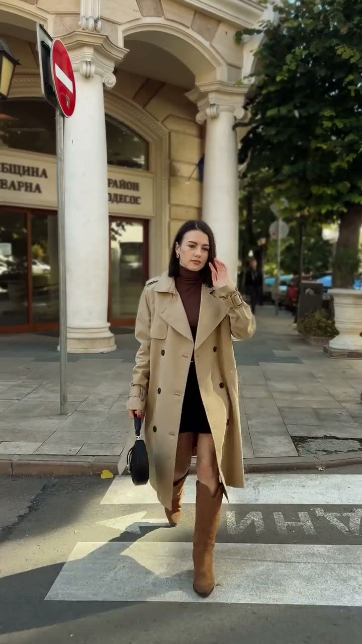 Mirela walking through a city street in a beige trench coat and boots for a fashion UGC video.